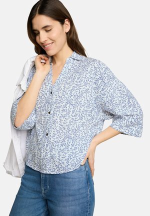 Femme portant une chemise bleue à fleurs boutonnée et un jean, tenant une veste blanche sur son épaule, souriant les yeux fermés.