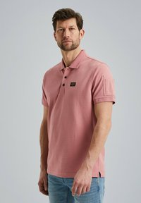 Light pink polo shirt with a collared neck, three-button placket, and a small logo patch. Short sleeves with a side slit. Casual fit.
