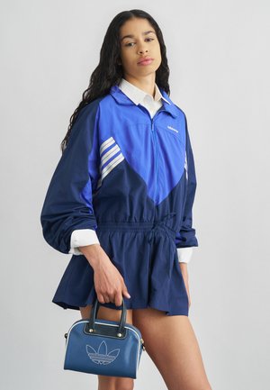 Žena oblečená v modrej Adidas bunde a sukni, drží malú modrú Adidas kabelku na jednofarebnom sivom pozadí.