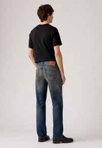 Dark jeans with a faded wash, featuring a brown leather patch at the back waist and a straight-leg cut. Worn with a black t-shirt and boots.