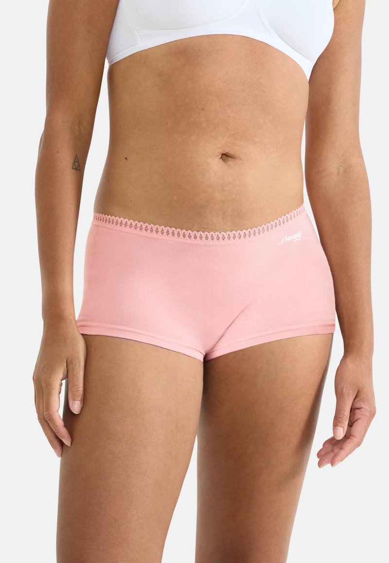 Γυναίκα που φοράει ανοιχτό ροζ εσώρουχα τύπου hipster και λευκό αθλητικό σουτιέν, στέκεται με τα χέρια χαλαρά στο πλάι.