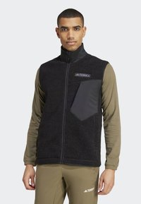 Schwarze Fleece-Weste mit hohem Kragen, Reißverschluss vorne und einer grauen Brusttasche. Getragen über einem hellgrünen Langarmshirt. Robustes und strukturiertes Material.