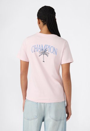 Vrouw met gevlochten haar, die een lichtroze T-shirt draagt met blauwe "CHAMPION" tekst en een afbeelding van een palmboom op de achterkant, gecombineerd met lichte spijkerbroek.