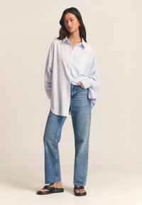 Camicia azzurro chiaro con bottoni e taschino, abbinata a jeans blu a vita alta e gamba dritta e sandali neri flat. Tessuto morbido e vestibilità comoda.
