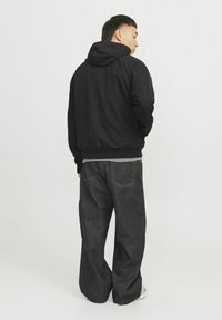 Schwarze Kapuzenjacke mit glatter Textur, kombiniert mit weit geschnittenen dunklen Denim-Jeans, die gelbe Nähte aufweisen.