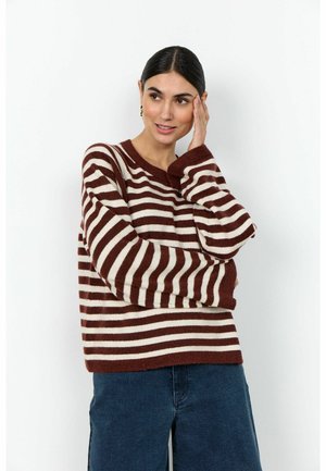 Sötét hajú nő arany fülbevalóval, barna-fehér csíkos pulóverben, az egyik kezét az arcához emeli, enyhén mosolyogva.