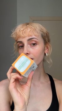 Boîte carrée orange vif et blanche pour la crème à la vitamine C de Mario Badescu, tenue dans une main, surface lisse avec des coins arrondis et une étiquette bleue.