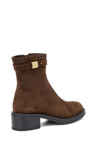 Stivaletto in suede marrone con punta arrotondata, tacco basso a blocco e una fascia decorativa con dettagli in fibbia dorata.