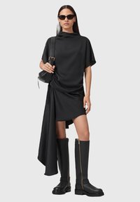 Abito asimmetrico nero con tessuto drappeggiato, stivali neri fino al ginocchio con zip dorata e una piccola borsa nera. La modella indossa occhiali da sole.