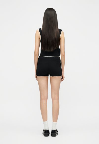 Femme aux longs cheveux noirs portant un haut noir sans manches et un short assorti avec une bordure en dentelle blanche, des chaussettes blanches et des chaussures noires, debout de dos.