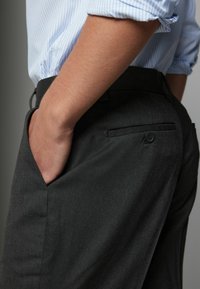 Pantalon ajusté gris foncé en tissu texturé, avec des poches latérales et une poche arrière avec fermeture à bouton, porté avec une chemise bleue rayée.