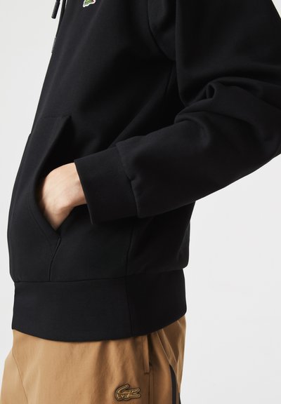 Sweatshirt noir avec une poche kangourou, des poignets côtelés et une ceinture. Présente un petit logo Lacoste sur la poitrine. Le matériau semble doux.