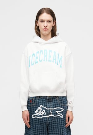 Νεαρό άτομο που φοράει λευκό φούτερ με κουκούλα με το κείμενο "ICECREAM" και μπλε παντελόνι με σχέδιο γαλοπούλας, με λευκό σχεδιασμό καρτούν γάτας.