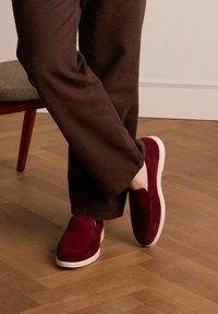 Burgundy slip-on shoes with a suede upper, featuring a white rubber sole. Worn with brown textured wide-leg trousers on a wooden floor.