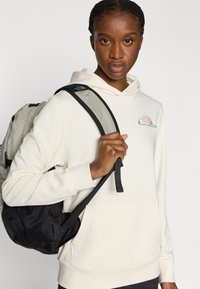 Μπλούζα με κουκούλα σε κρεμ χρώμα με μπροστινή τσέπη, που φέρει μικρό πολύχρωμο λογότυπο. Μαύρο και γκρι σακίδιο πλάτης με διακοσμητικά από δίχτυ, φορεμένο πάνω σε έναν ώμο.