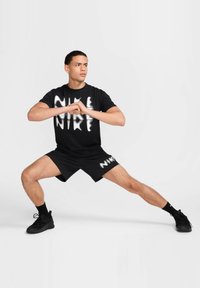 Zwarte Nike T-shirt met witte graphic, zwarte shorts met het Nike-logo, zwarte sokken en zwarte sportschoenen. Model in een brede houding.