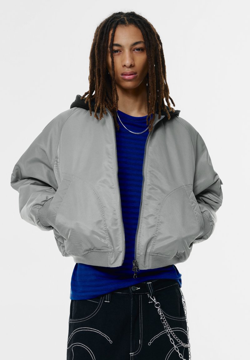 Grijze bomberjack met capuchon, voorzien van zijzakken, geribbelde manchetten en een gladde textuur, gedragen over een blauw gestreept overhemd.