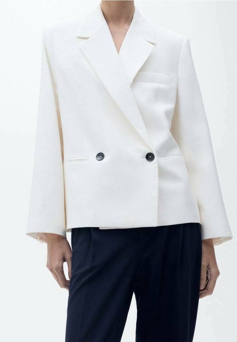 Veste blanche à double boutonnage avec des boutons noirs, portée avec un pantalon foncé, montrant le haut du corps et les bras sur un fond uni.