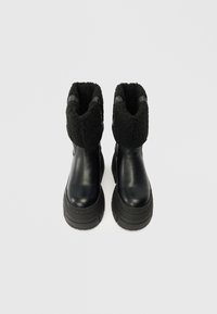 Botas de tornozelo em couro preto com uma gola de lã preta felpuda e uma sola grossa texturizada com "BUFFALO" estampado na lateral.