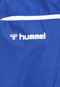 Blå sporttyg med texturerad yta, med en vit horisontell rand och den vita "hummel"-logotypen, accentuerad av en bi-grafik.