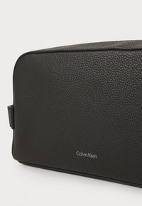 Black textured leather toiletry bag with Calvin Klein logo in silver lettering on front, rectangular shape with zipper closure.