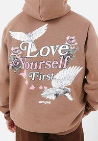 Brun hoodie med grafisk design och texten "Älska dig själv först", fåglar, blommiga accenter och fjärils-siluetter, tillverkad av mjukt tyg.