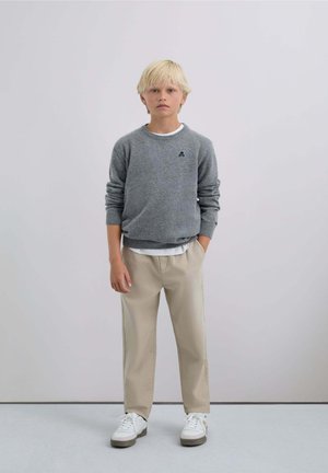 Suéter de punto gris con un pequeño logo, superpuesto a una camisa blanca; pantalones beige con rayas laterales; zapatillas blancas. Diseño simple y contemporáneo.