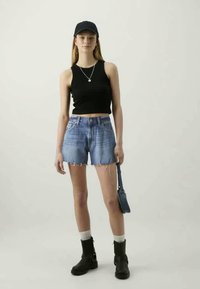 Νεαρή γυναίκα φορώντας μαύρο crop top, τζιν σορτσάκια, μαύρες μπότες, μαύρο καπέλο, κρατώντας μια μικρή μπλε τσάντα μπροστά σε απλό φόντο.