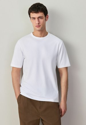 Mladý muž stojaci opretý o svetlú stenu, oblečený v jednoduchej bielej krátkosleevej tričku a hnedej nohaviciach, s jednou rukou v vrecku.