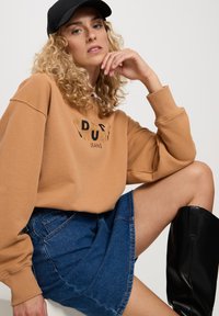 Brązowa bluza z czarnym napisem "RUSH JEANS", w połączeniu z niebieską dżinsową spódnicą i czarnymi botkami do kolan. Modelka nosi czapkę w kolorze czarnym.