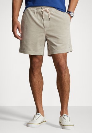 Man die beige corduroy shorts met trekkoord, een blauw shirt, witte sneakers en een horloge draagt, staand met één hand in de zak.