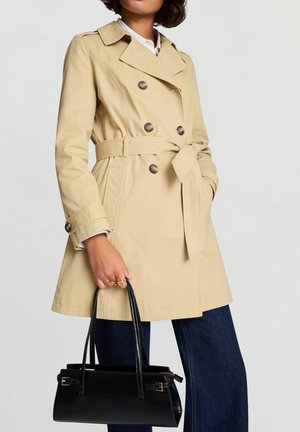 Trench coat beige doppiopetto con cintura, caratterizzato da bottoni rotondi marroni. Abbinato a una borsa nera e pantaloni di jeans scuri.