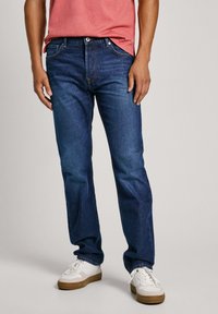 Vaqueros de mezclilla azul oscuro con corte recto, con un diseño clásico de cinco bolsillos y costura blanca. Combinados con zapatillas blancas.