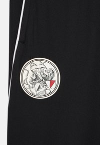 Svarta joggers med en rund vit patch som har en intrikat illustration av två huvuden och texten "AJAX", accentuerad med en röd skölddetalj.