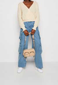 Cream knitted wrap sweater with a V-neck, paired with light blue wide-leg cargo jeans, and a small beige bag with two zippered pockets.