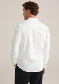 Uomo con capelli ricci che indossa una camicia bianca a maniche lunghe e pantaloni neri, in piedi di spalle contro uno sfondo semplice.