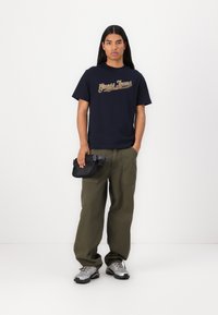T-shirt navy con logo dorato "Guess Jeans", pantaloni larghi color oliva e sneakers grigie. Tiene una piccola borsa nera con una tracolla. Design semplice.