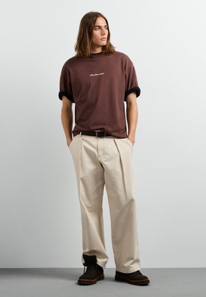 Junger Mann mit schulterlangen Haaren trägt ein braunes T-Shirt, beige weite Hose, schwarzen Gürtel und schwarze Stiefel, steht mit den Händen in den Taschen.