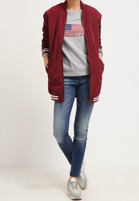 Maroon bomberjakke med hvid-stribede manchetter, grå sweatshirt med amerikansk flag print, blå skinny jeans og grå sneakers.