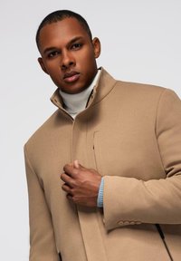 Beige maßgeschneiderter Mantel mit hohem Kragen, strukturiertem Stoff und einer Seitentasche, getragen über einem hellblauen gerippten Pullover.