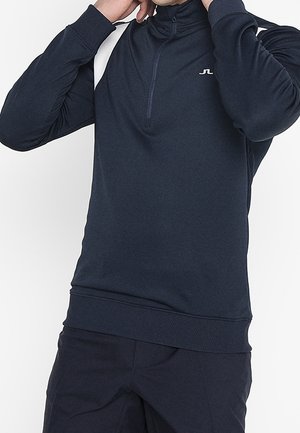 Ein Mann richtet den Kragen eines marineblauen Langarmpullovers mit Viertel-Reißverschluss und kleinem weißen Logo auf der Brust.