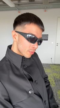 Joven con cabello corto y oscuro, lleva gafas de sol negras y un abrigo oscuro abotonado, en un entorno de oficina interior.