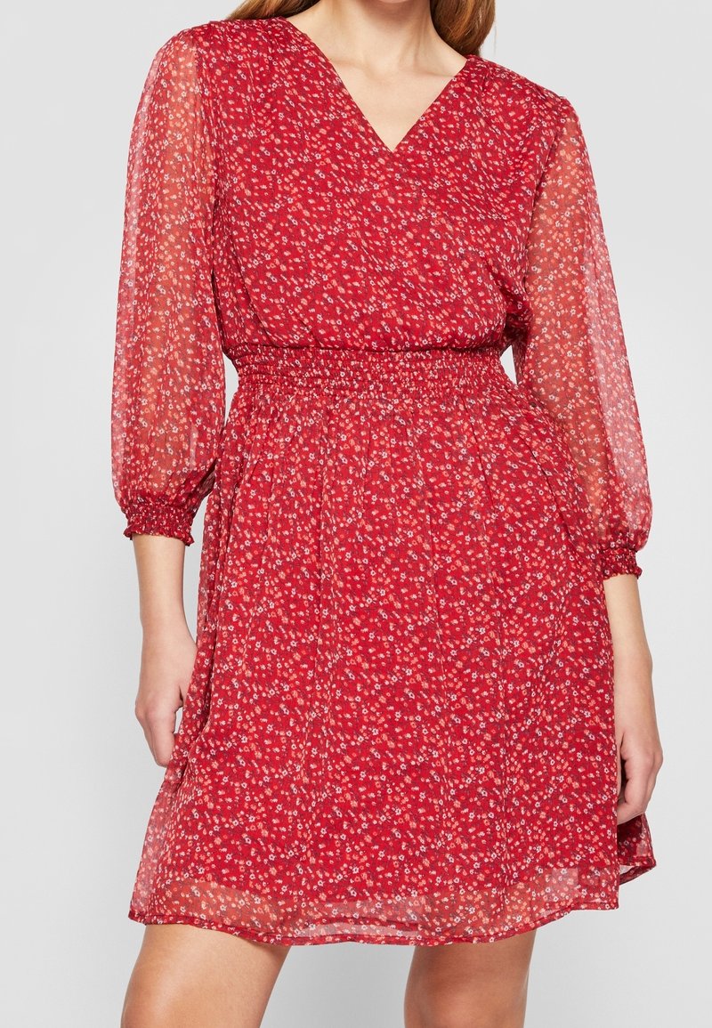 Robe fleurie rouge avec un décolleté en V, des manches longues transparentes et une taille cintrée. Elle a un tissu léger et texturé ainsi qu'une jupe longueur genou.