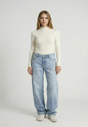 Kobieta stojąca przodem, ubrana w biały prążkowany długi rękaw, jasnoniebieskie szerokie jeansy i białe trampki na jednolitym tle.