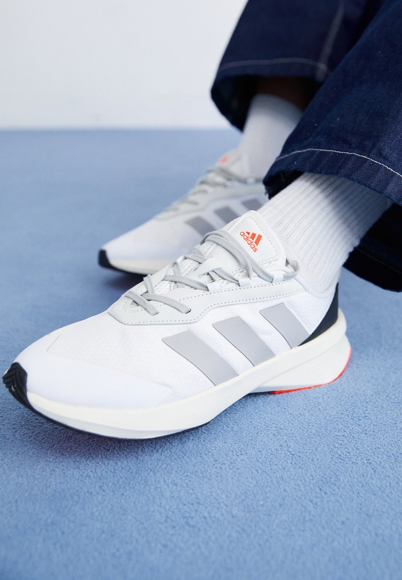 Scarpe da ginnastica bianche con accenti grigi, caratterizzate da un design a tre strisce, chiusura con lacci, tessuto texturizzato e suola nera e rossa a contrasto.