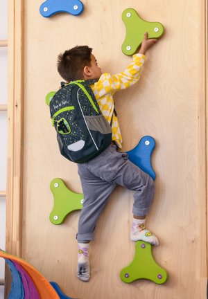 Climbing wall with colorful holds in green and blue shapes. A child in a yellow checkered shirt and gray pants is reaching for a hold.