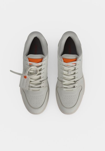 Ζευγάρι λευκά sneakers χαμηλού τύπου με πορτοκαλί λεπτομέρεια στη γλώσσα, λευκά κορδόνια και μια μικρή ετικέτα προσαρτημένη στα κορδόνια του αριστερού παπουτσιού, από πάνω.