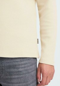 Śmietankowa, dzianinowa koszula z długim rękawem o teksturowanej tkaninie; posiada metkę w dolnej części i jest zestawiona z szarymi spodniami o gładkiej fakturze.