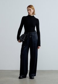 Melna garām piedurknēm augša un plaša griezuma melnas bikses ar aukliņu jostasvietā, kopā ar teksturētu melnu roku somu un melnām kurpēm.