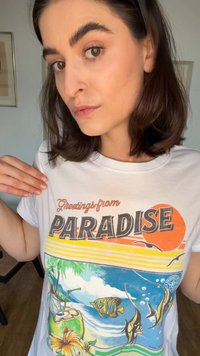 Jeune femme aux cheveux bruns pointe un t-shirt blanc avec le texte « Greetings from Paradise » et une illustration de plage tropicale avec un poisson.